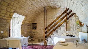 Una habitación con paredes de piedra, una escalera de madera, una mesa con mantel blanco y un suelo de baldosas rojas.