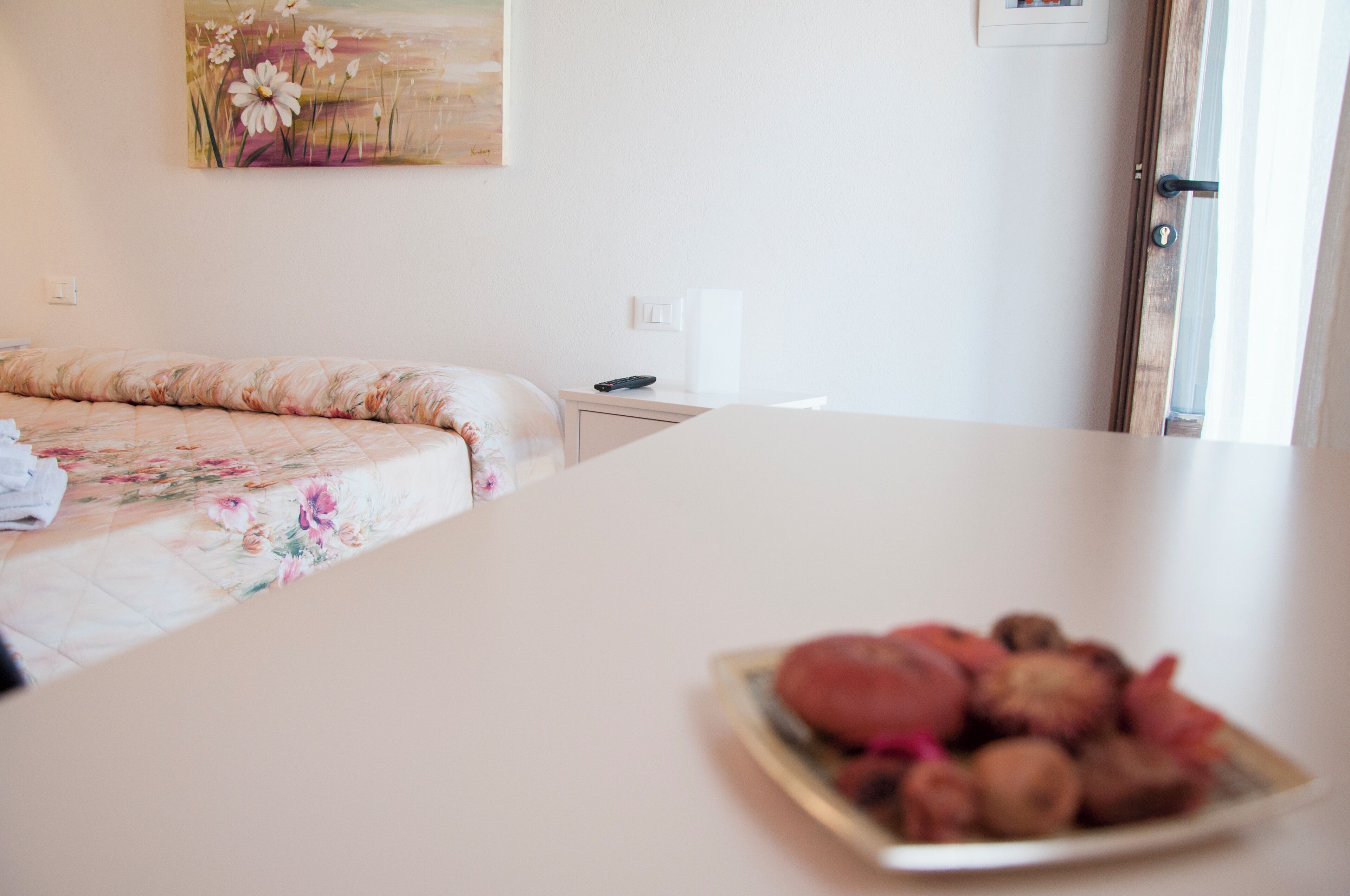 A hotel room with a bed, a bedside table, a plate of nuts, a painting on the wall, and a door.