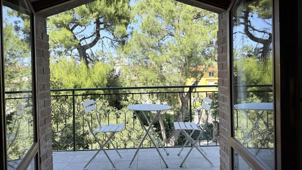 Ein Balkon mit Blick auf Bäume und ein Gebäude. Zwei weiße Tische und vier Stühle sind vorhanden.