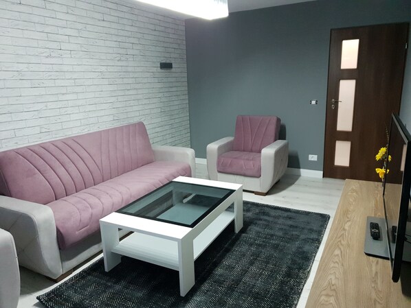 Un salon avec un canapé blanc, un fauteuil rose, une table basse en verre et un plancher en bois.