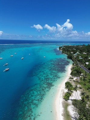 Vista aérea de una playa tropical con aguas turquesas cristalinas, barcos y una carretera que conduce a una zona residencial con palmeras.