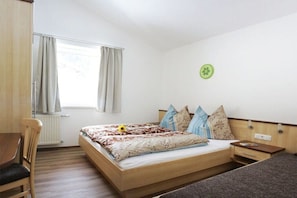 Ein Schlafzimmer mit einem Bett, einem Sessel, einem Nachttisch, einem Fenster mit Vorhängen und einer Wanduhr.