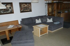 Ein Wohnzimmer mit grauem Sofa, einem Holztisch und einer Bank. An der Wand hängen gerahmte Bilder und im Hintergrund ist eine Küchenzeile zu sehen.