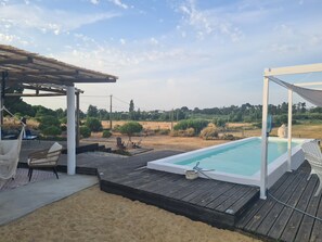 Een zwembad met een houten terras, een overdekte zithoek en uitzicht op het omliggende landschap.