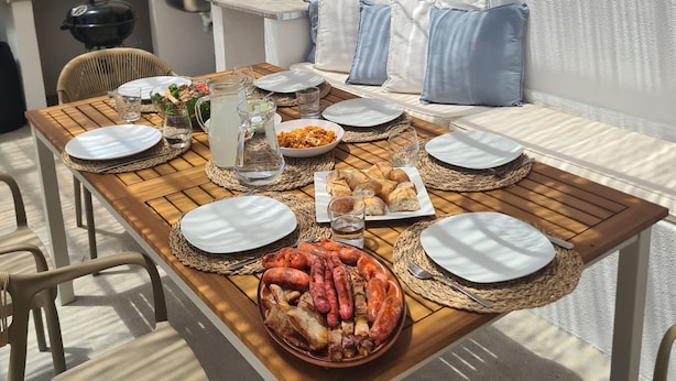An outdoor dining area with wicker pendant lights, a wooden table set with plates, glasses, and food, and a view of the kitchen and living area in the background. - Farmhouse CASA AYO (Santiago do Cacém)