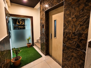 Zona de recepción de un hotel con puerta de cristal, mostrador de recepción, planta en maceta, área pequeña de césped artificial y pared de mármol.