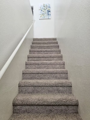 Escada revestida com carpete e corrimão, conduzindo a um quarto com parede branca e uma obra de arte floral.
