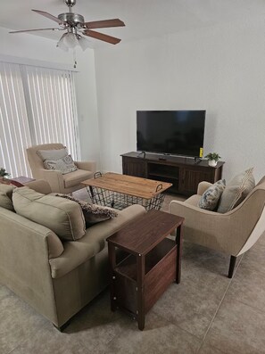 Ein Wohnzimmer mit Sofa, Sessel, Couchtisch aus Holz, TV-Möbel und Deckenventilator.