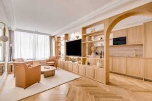 Ein modernes Wohnzimmer mit Sofa, Couchtisch und Fernseher, verbunden mit einer Küchenzeile mit Holzschränken und einem Fischgrätparkett.