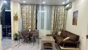 Una sala de estar con un sofá marrón, una mesa de centro de madera, una mesa de comedor con sillas y cortinas estampadas.