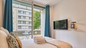 Ett hotellrum med en säng, en tv, utsikt över en byggnad och en balkong med ett bord och stolar.