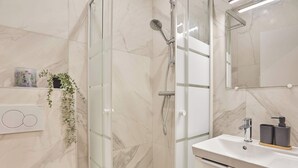 Ein modernes Badezimmer mit einer Duschkabine aus Glas, einem weißen Waschbecken mit schwarzem Seifenspender und einer Wand mit Marmorplatten.