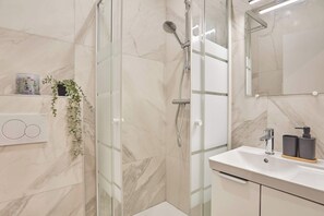 Ein modernes Badezimmer mit einer Duschkabine aus Glas, einem weißen Waschbecken mit schwarzem Seifenspender und einer Wand mit Marmorplatten.