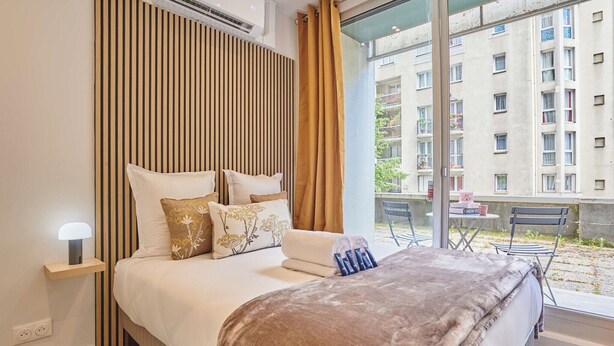 Ein Hotelzimmer mit Bett, Nachttisch, Lampe, einem Fenster mit Vorhängen und Blick auf ein Gebäude.