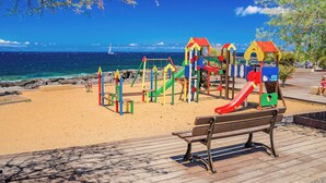 Un parque infantil en la playa con toboganes y estructuras para escalar, un banco de madera y una vista despejada del océano.