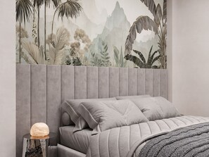 Ein Schlafzimmer mit grauem, gepolsterten Kopfteil, einem Bett mit grauer Bettwäsche und einer Wandmalerei von tropischem Grünwerk.
