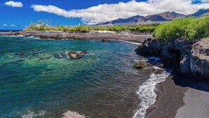 Una playa con aguas turquesas cristalinas, costa rocosa y montañas a lo lejos.
