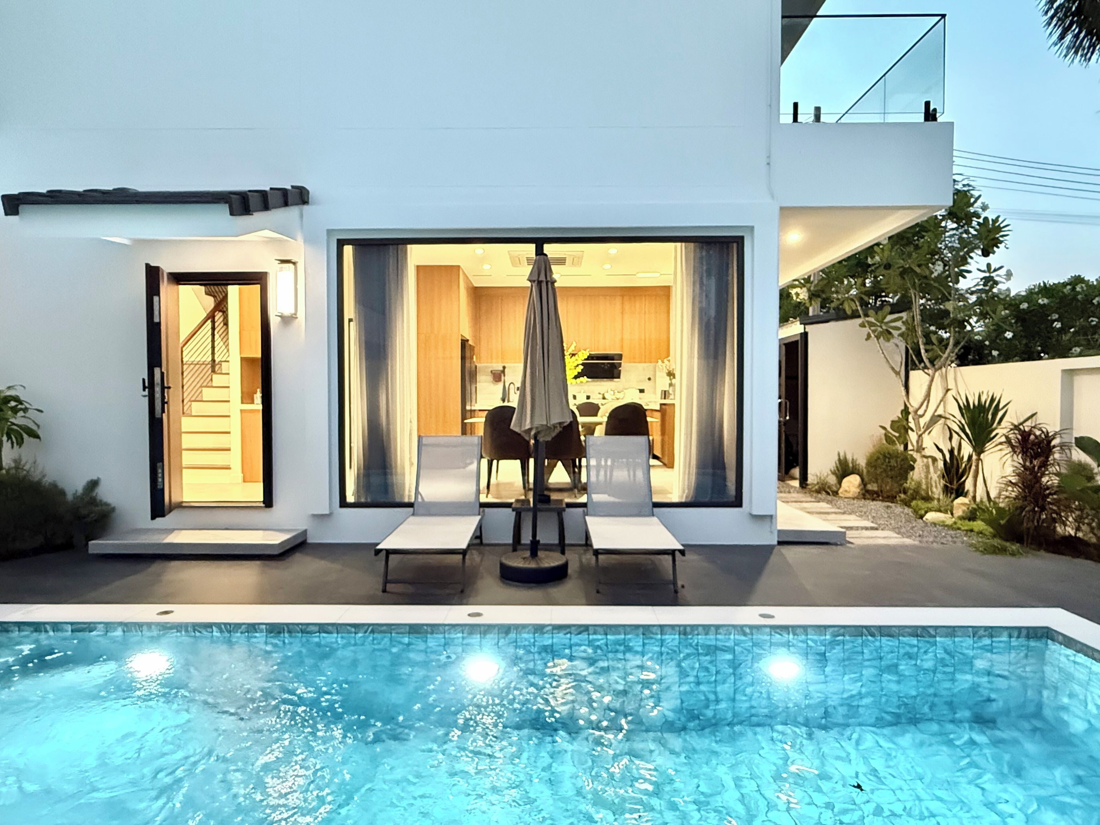 Ein modernes Haus mit Swimmingpool, Essbereich und sichtbarer Küche hinter großen Glasüren.