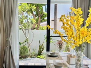 Ein Zimmer mit Blick auf einen Garten, ein Tisch mit einer Vase voller gelber Blumen und ein Fenster mit Vorhängen.