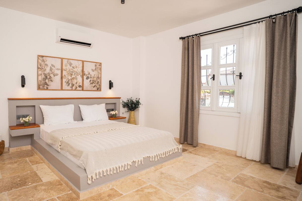 A bedroom with a bed, bedside tables, a wall-mounted air conditioner, a window with curtains, and a decorative wall piece.