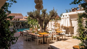 A poolside area with lounge chairs and tables, a stone wall, and a view of a building with a tiled roof. - Molino Hotel (Çeşme)