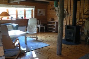 En stuga med träpanel på insidan, inredd med en soffa, stolar, ett bord, en öppen spis och ett fönster med utsikt.