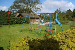 Eine Schaukelanlage in einer grünen Wiese, im Hintergrund ein Haus und Bäume.