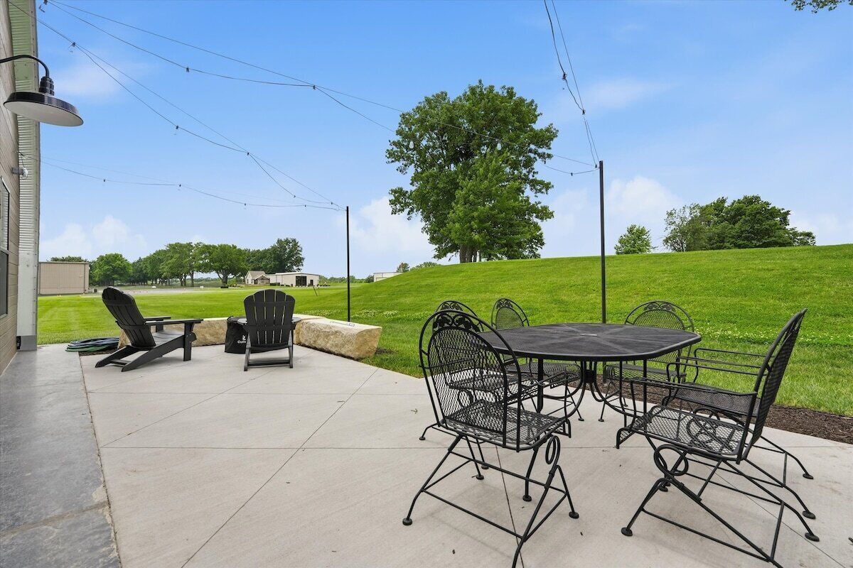An outdoor patio with a table and chairs, a lamp post, and a view of a grassy hill with trees.