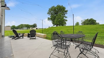 An outdoor patio with a table and chairs, a lamp post, and a view of a grassy hill with trees.