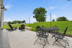 An outdoor patio with a table and chairs, a lamp post, and a view of a grassy hill with trees.