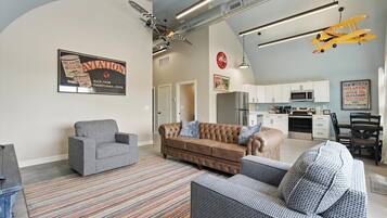 Un salon avec un canapé, des fauteuils et un tapis à motifs. On aperçoit à l’arrière-plan un coin cuisine avec des placards et des appareils électroménagers.