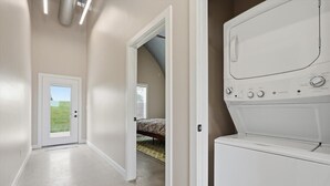 Une buanderie équipée d’un lave-linge et d’un sèche-linge, une porte donnant sur une chambre et un couloir avec une porte menant à l’extérieur.