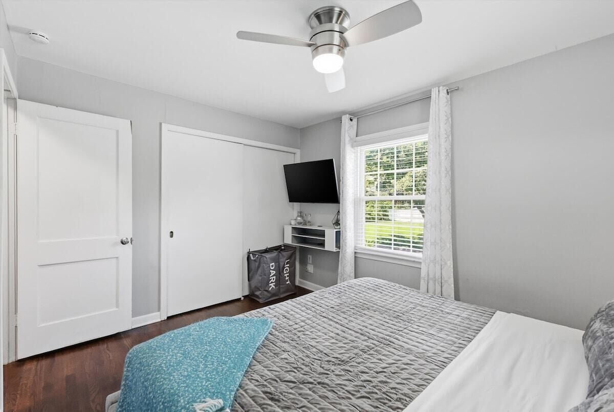 A bedroom with a bed, a ceiling fan, a television, a window with curtains, and a door.