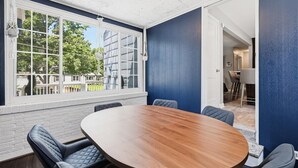 A conference room with a large wooden table and four chairs, a large window with a view of buildings, and a door leading to another room. - Waldo Blue Haven KC Home With Pergola Deck (Kansas City)