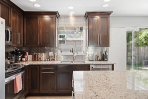 A modern kitchen with dark wood cabinets, a granite countertop, stainless steel appliances, and a large window. - Waldo Blue Haven KC Home With Pergola Deck (Kansas City)