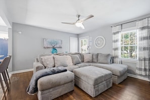 A modern living room with a sectional sofa, a ceiling fan, and a large window with curtains.