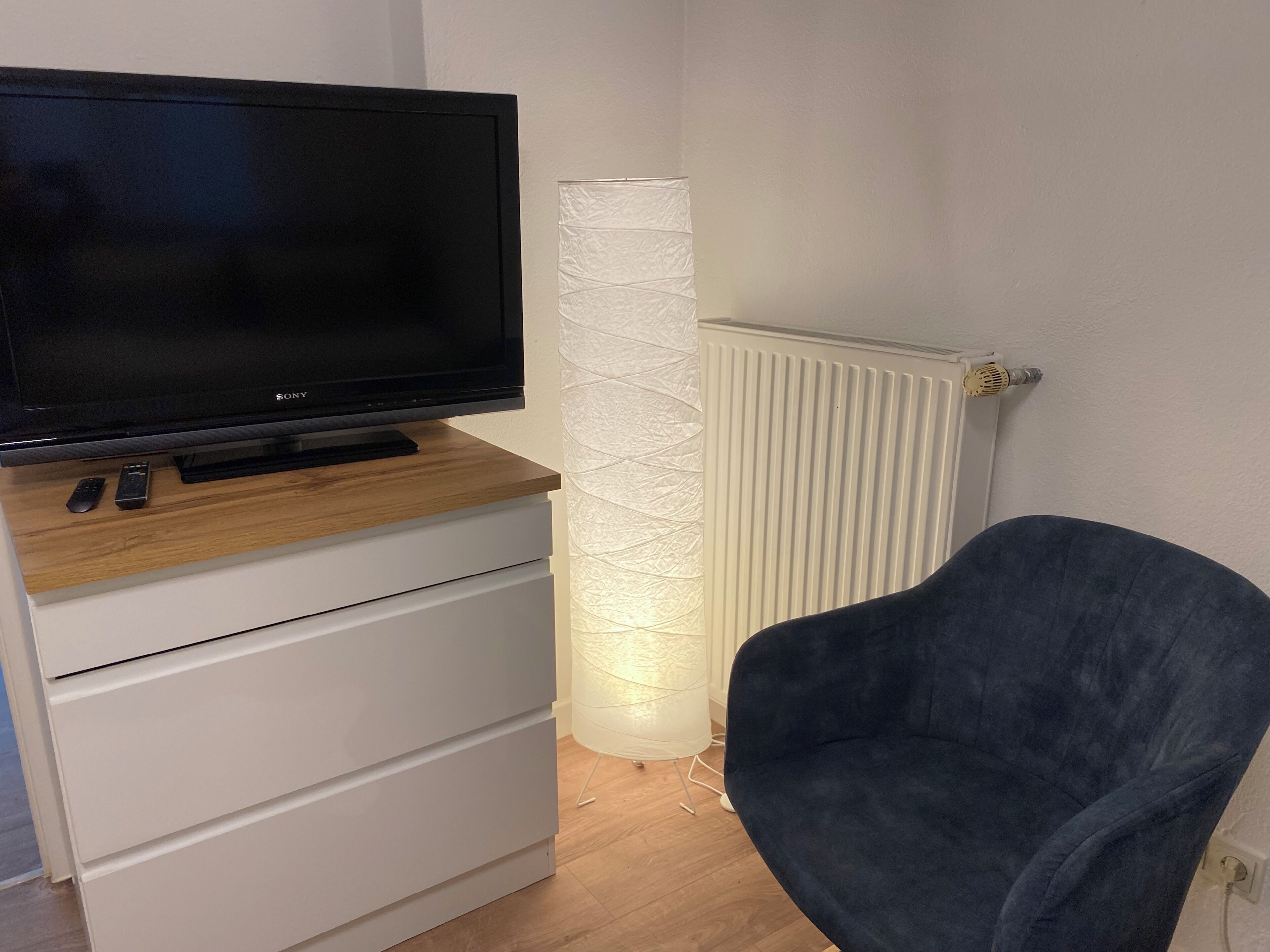 Ein Flachbildfernseher auf einem Holzregal, eine weiße Schubladeneinheit, eine hohe weiße Lampe und ein dunkelblauer Sessel.