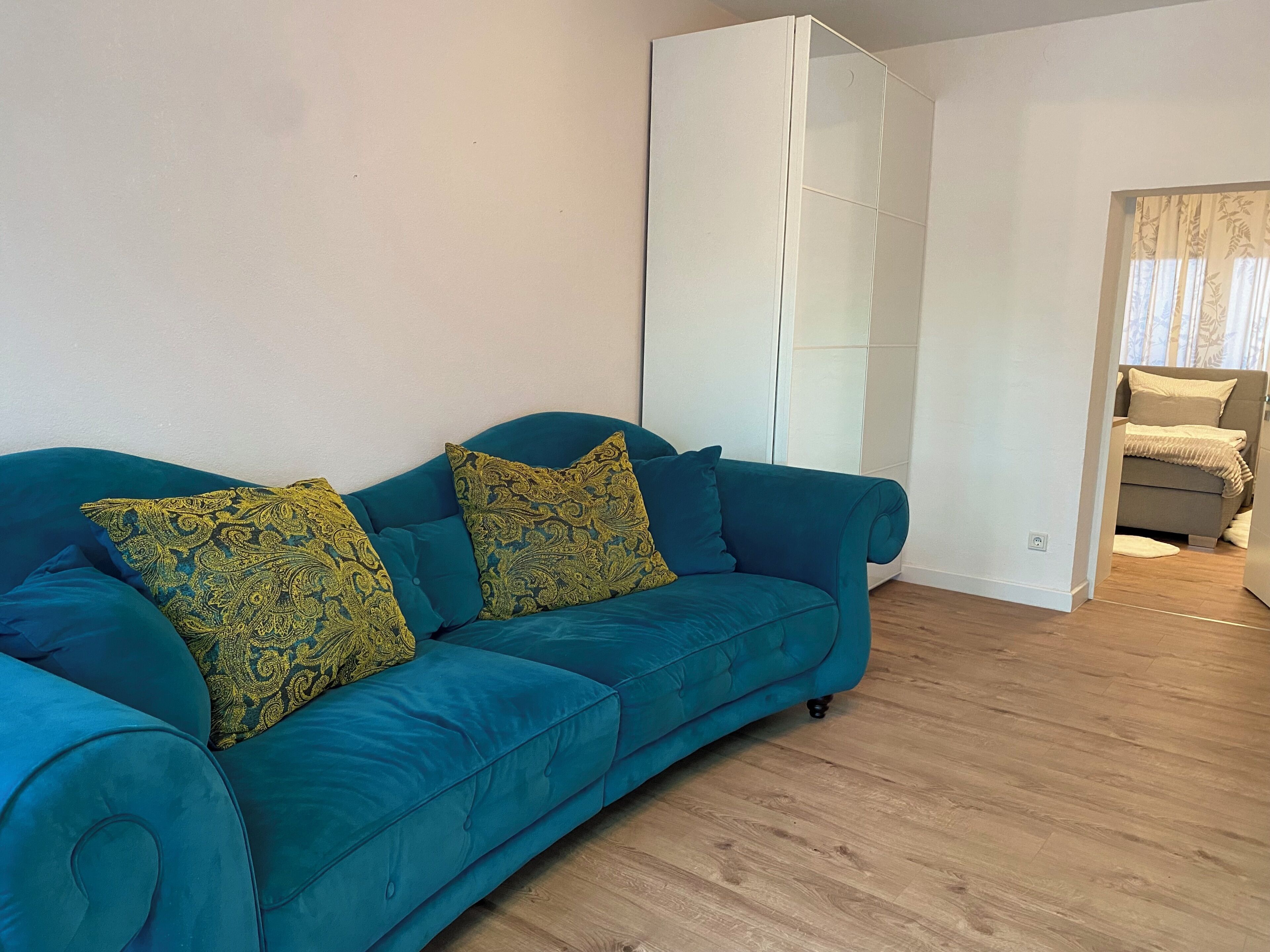 A teal sofa with patterned cushions, a white wardrobe, and a wooden floor.