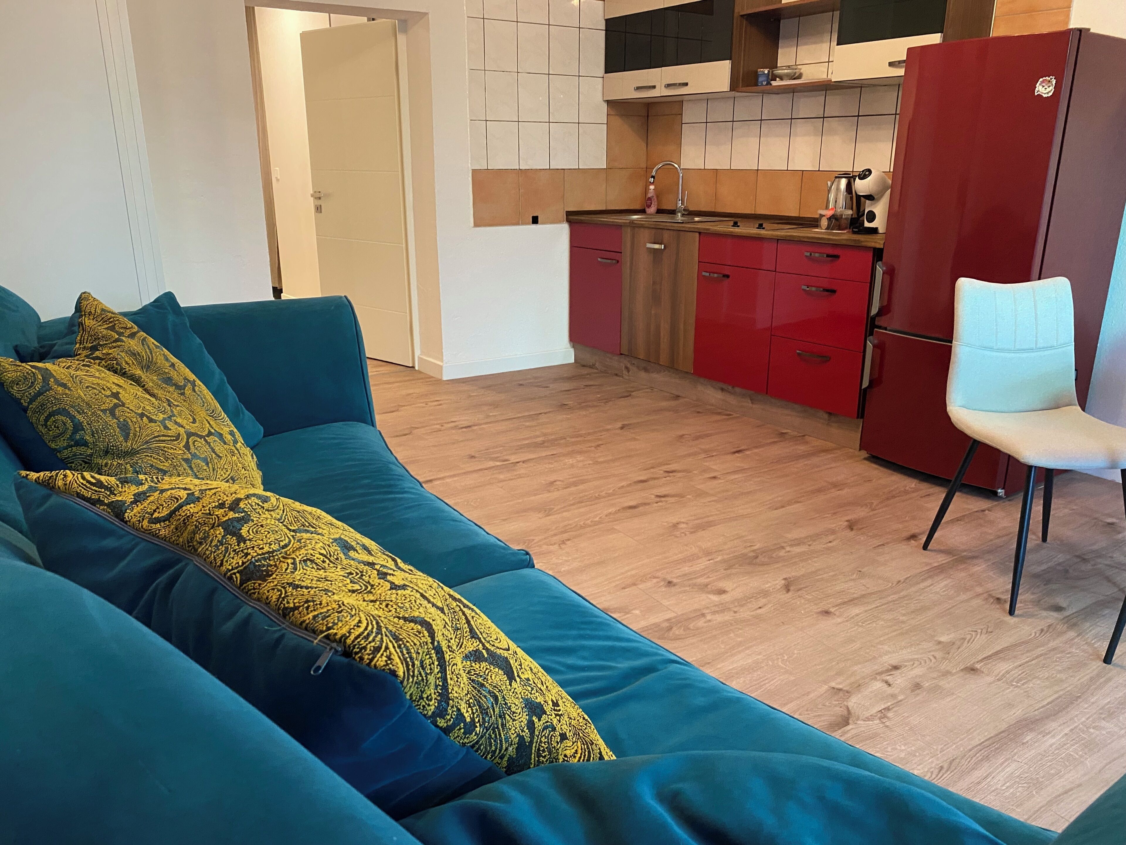 A living room with a teal sofa, a white chair, and a kitchen area with red cabinets and a red refrigerator.