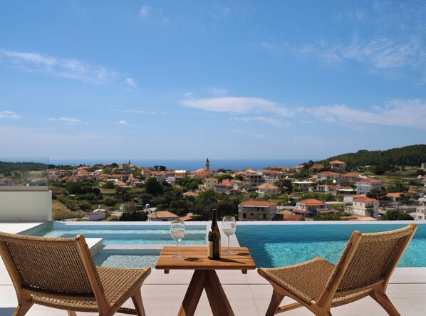A poolside view with two wicker chairs, a wooden table, and a bottle of wine.