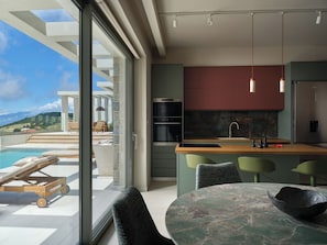 A modern kitchen with a large island, a built-in oven, and a view of a pool and mountains. - Casa Blanca Villa, Pool, SeaViews & Wine Cellar (Zakynthos)