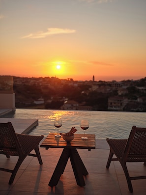 Une vue sur le coucher de soleil depuis une terrasse, avec deux verres de vin rouge et un bol de gressins sur une table en bois.