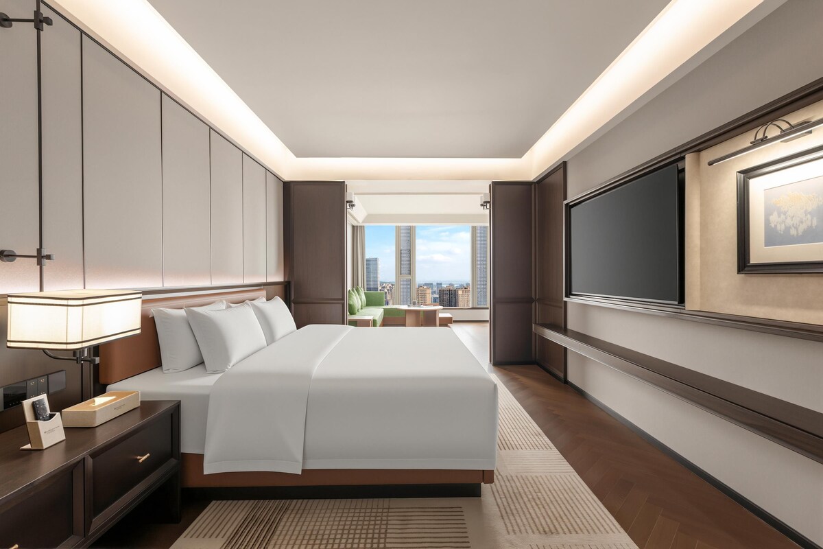 Quarto de hotel moderno com uma cama grande, TV de tela plana na parede e vista para a cidade pela janela.