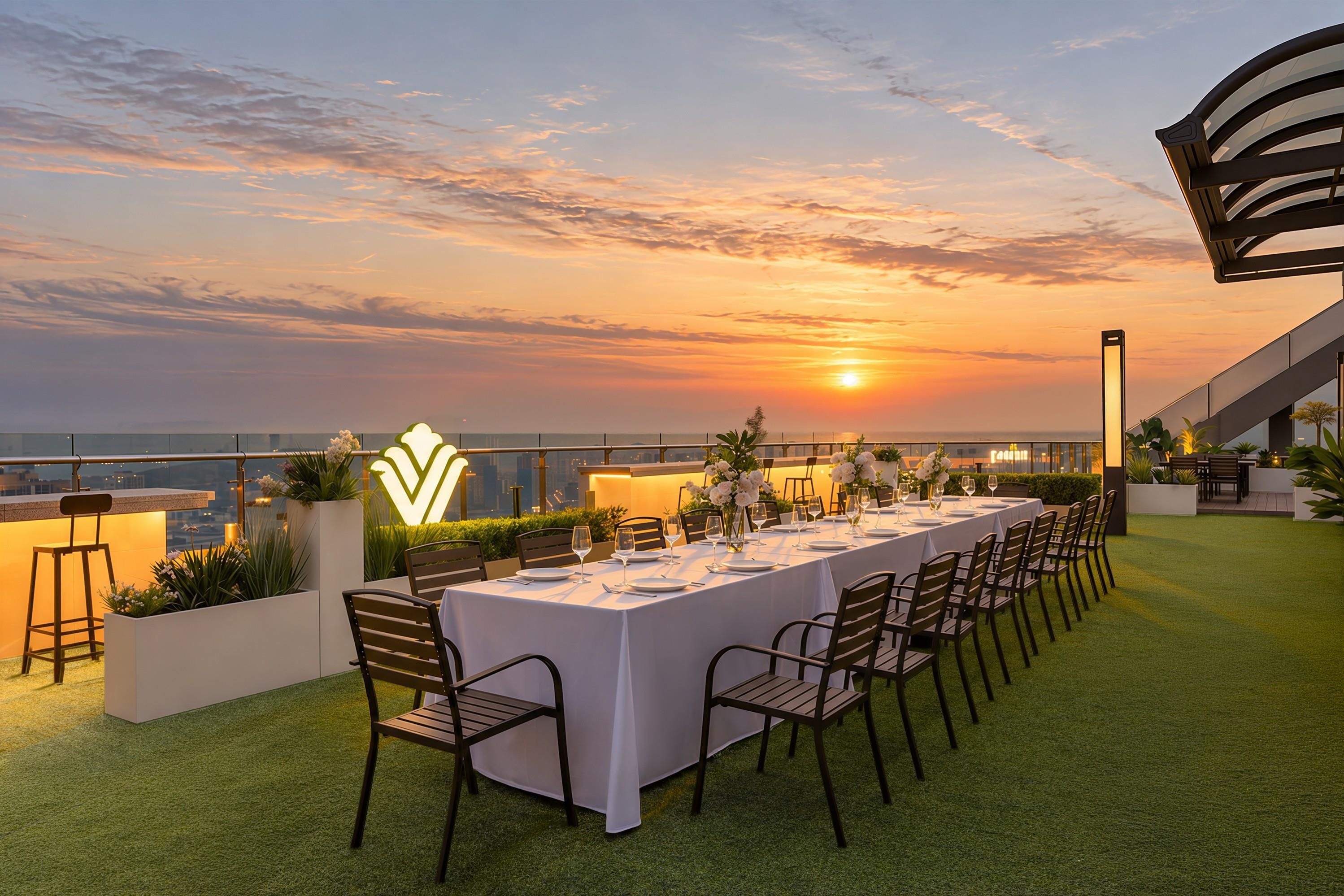 Un área de comedor al aire libre con vista al atardecer, una mesa larga preparada para una comida y una estructura arquitectónica moderna al fondo.