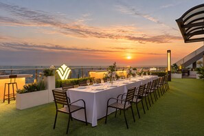 Uma área de refeições ao ar livre com vista para o pôr do sol, uma mesa comprida posta para uma refeição e uma estrutura arquitetônica moderna ao fundo.