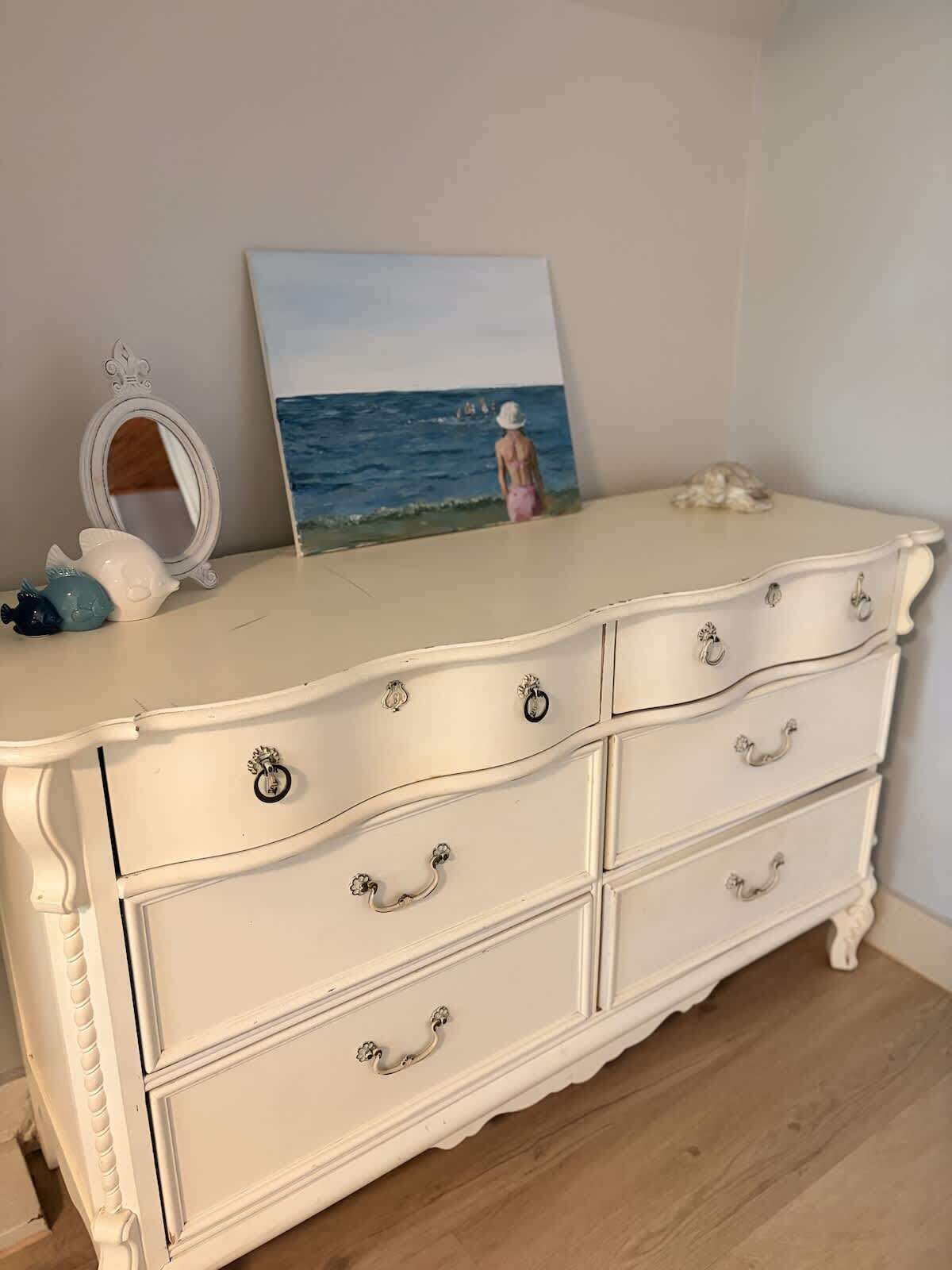 A white dresser with ornate handles, a small mirror, and a painting of a beach scene.