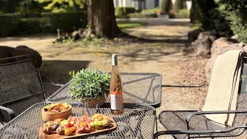 Un espace extérieur aménagé pour prendre les repas, avec des chaises en métal, une table où sont disposés de la nourriture et une bouteille, le tout entouré d’arbres et avec un bâtiment en arrière-plan.