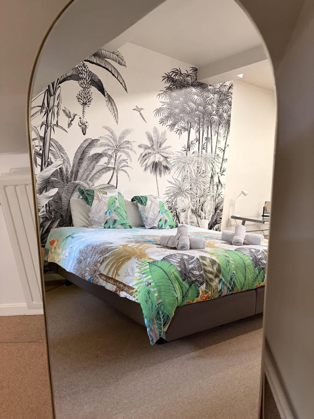 Un dormitorio con una cama, papel tapiz de temática tropical y un espejo que refleja la habitación.