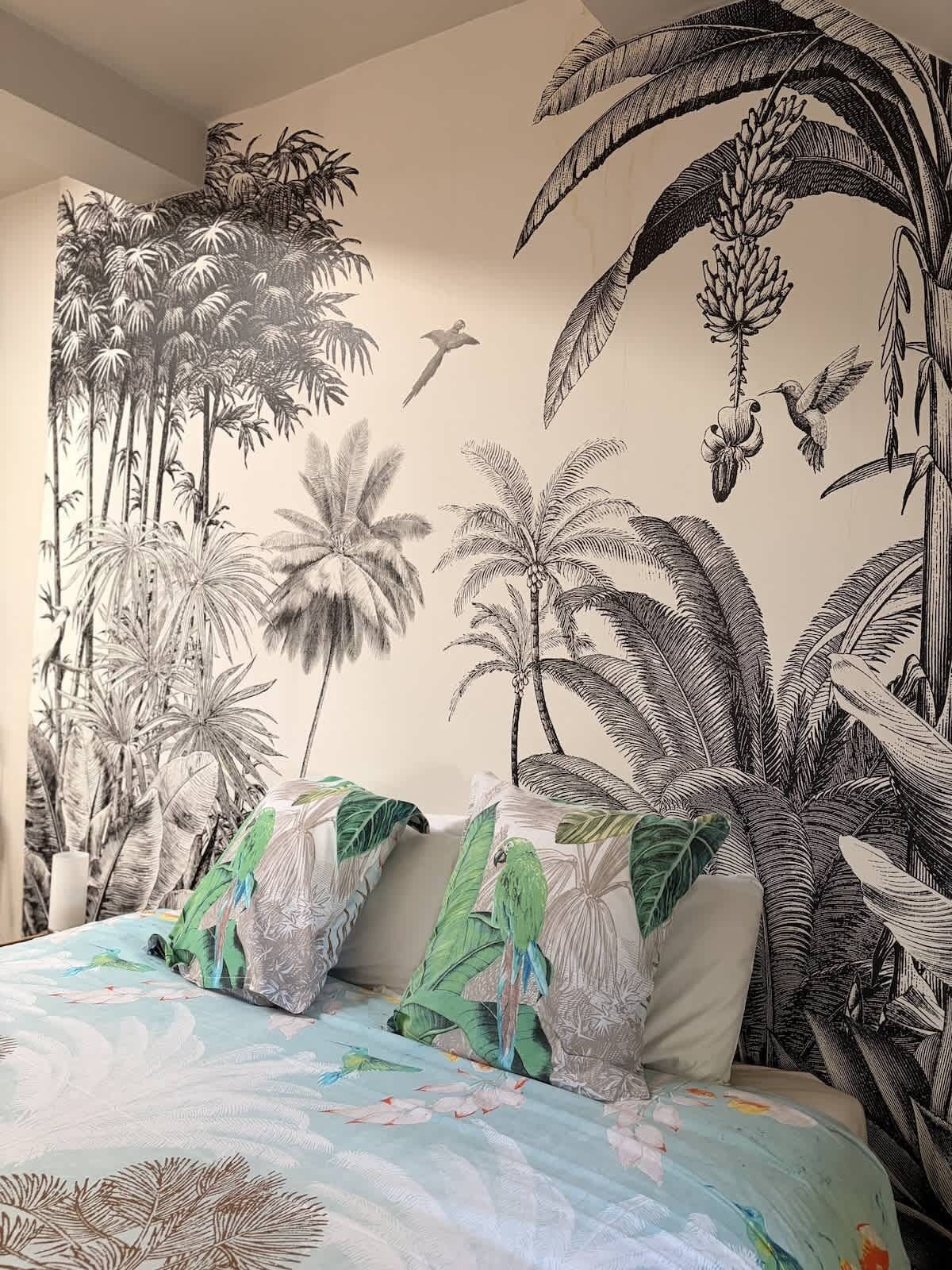 Une chambre avec une fresque représentant des plantes et des oiseaux tropicaux, un lit avec une literie aux couleurs vives et un oreiller orné d’un motif d’oiseau.
