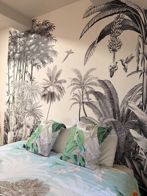 Ein Schlafzimmer mit einer Wandmalerei von tropischen Pflanzen und Vögeln, einem Bett mit farbenfroher Bettwäsche und einem Kissen mit Vogelmotiv.